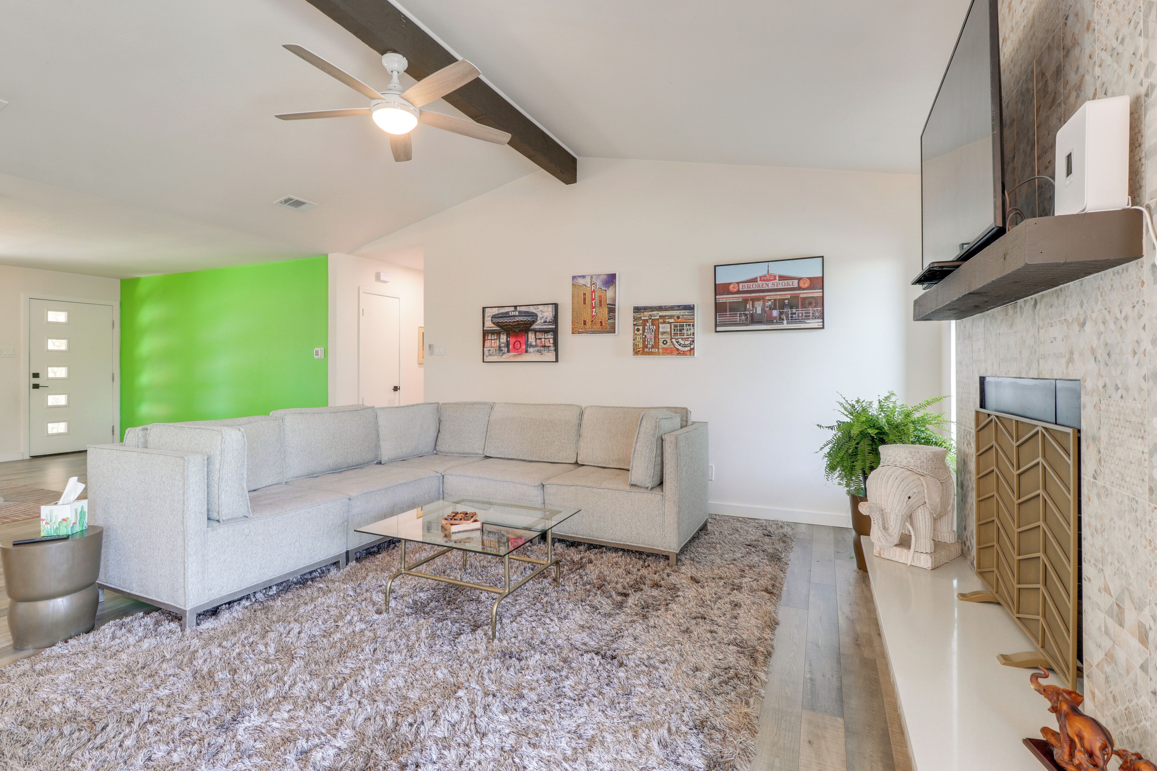 Uma sala de estar moderna com um sofá seccional, uma mesa de centro de vidro, um ventilador de teto e uma TV de tela plana fixada na parede.