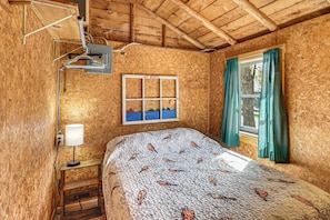 A room with a wooden ceiling, a bed with a patterned bedspread, a window with curtains, and a wall-mounted shelf with a lamp.