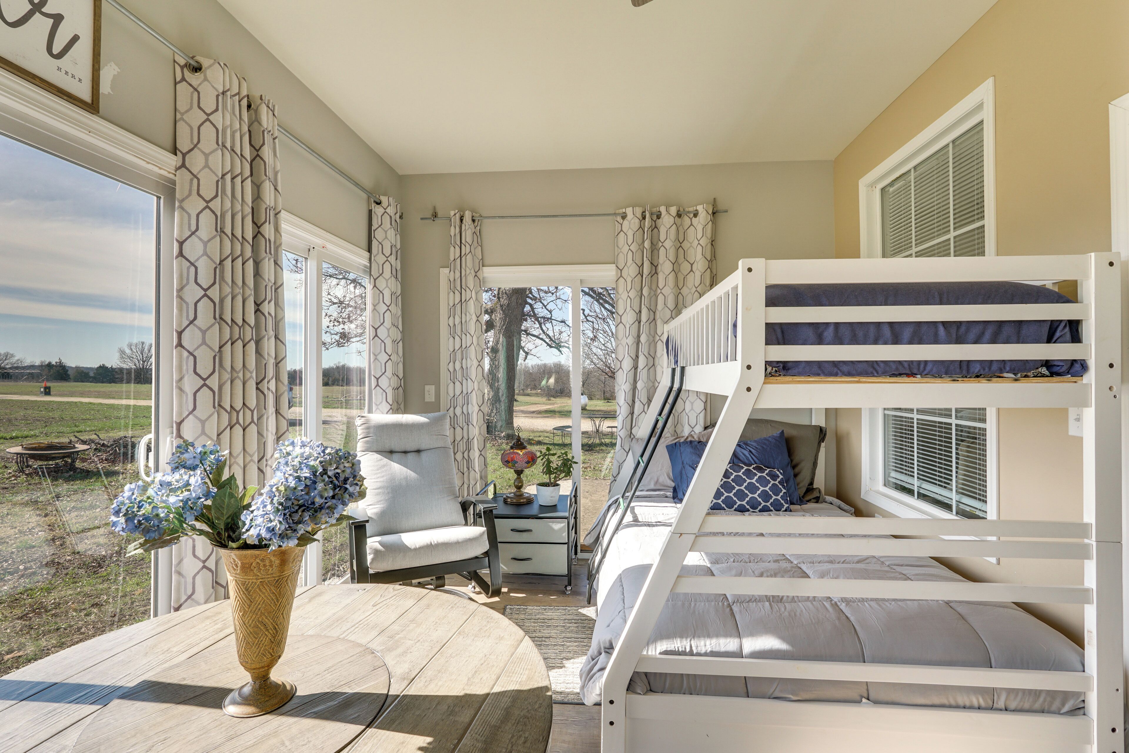 A room with a bunk bed, a chair, a small table with a vase, and a window with curtains.
