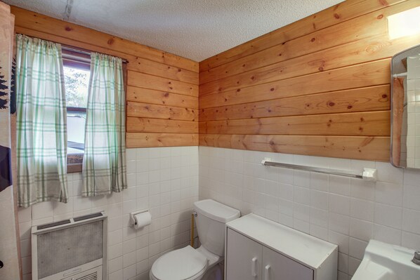 Un baño con paredes de paneles de madera, un inodoro, un lavabo y una ventana con cortinas.