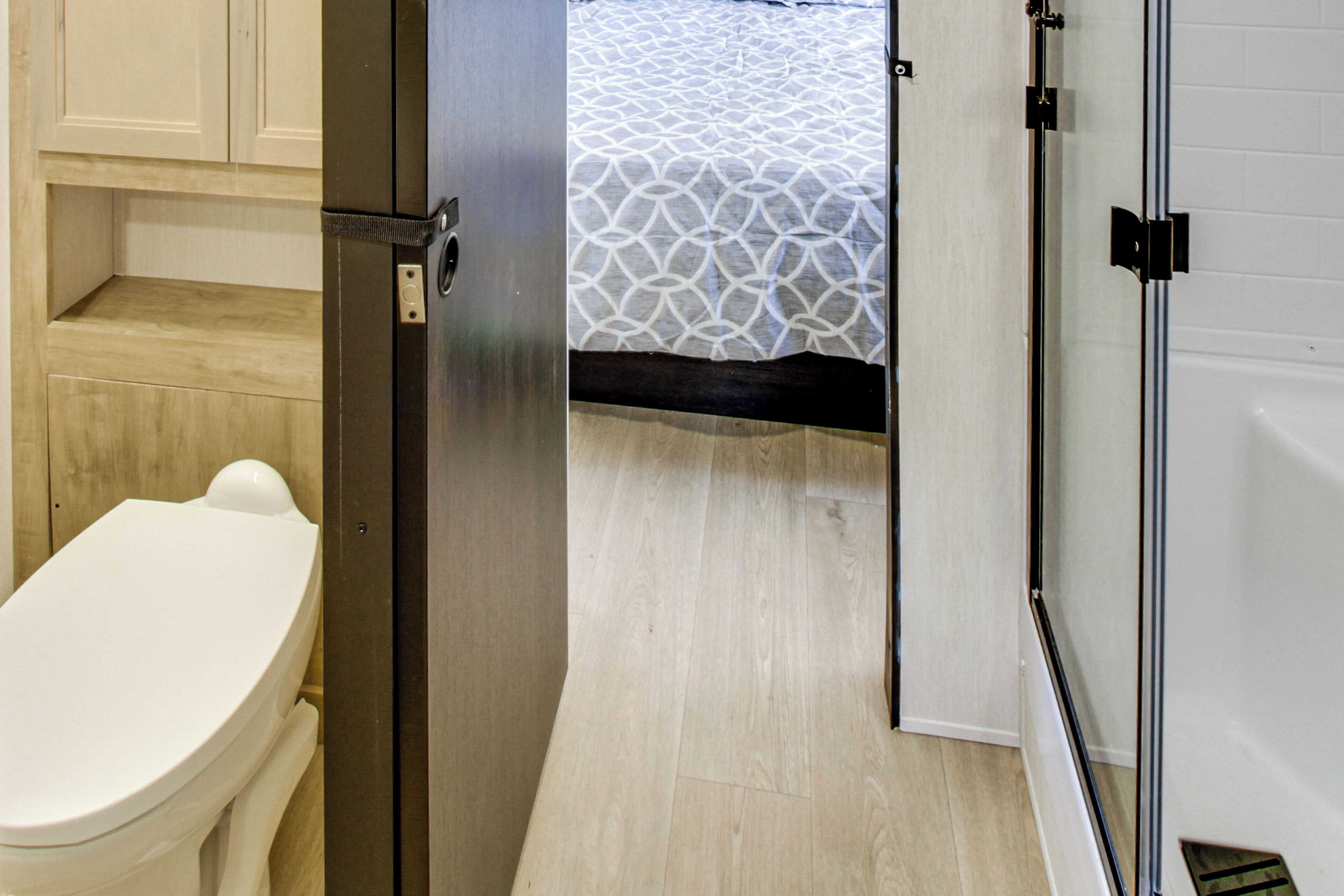 A bathroom with a toilet, a shower enclosure, and a patterned bed visible through an open door.