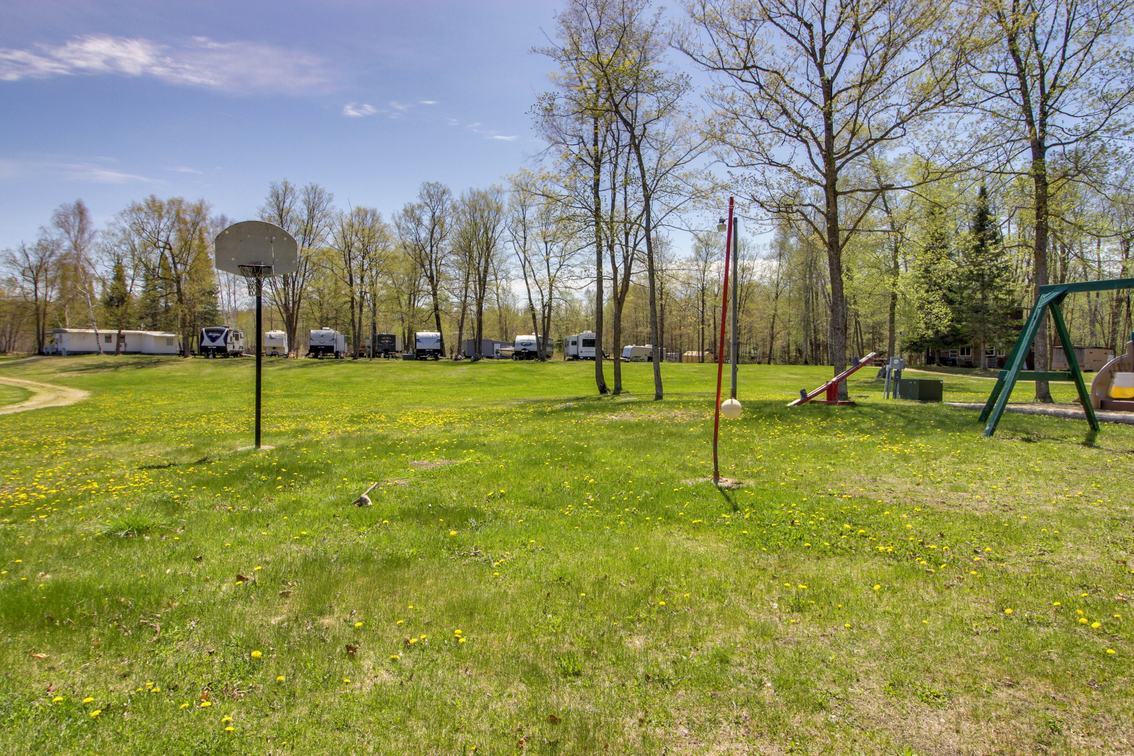 Um campo gramado com uma cesta de basquete, equipamentos de recreação infantil e trailers de recreação ao fundo.