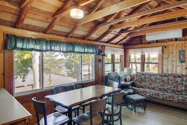 A wooden cabin interior with a dining table, chairs, a sofa, and large windows.