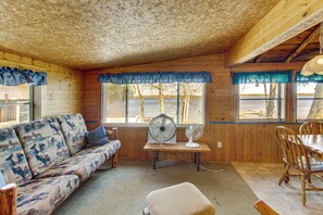 Interior de una cabaña de madera con un sofá, mesa de comedor, sillas y un ventilador.