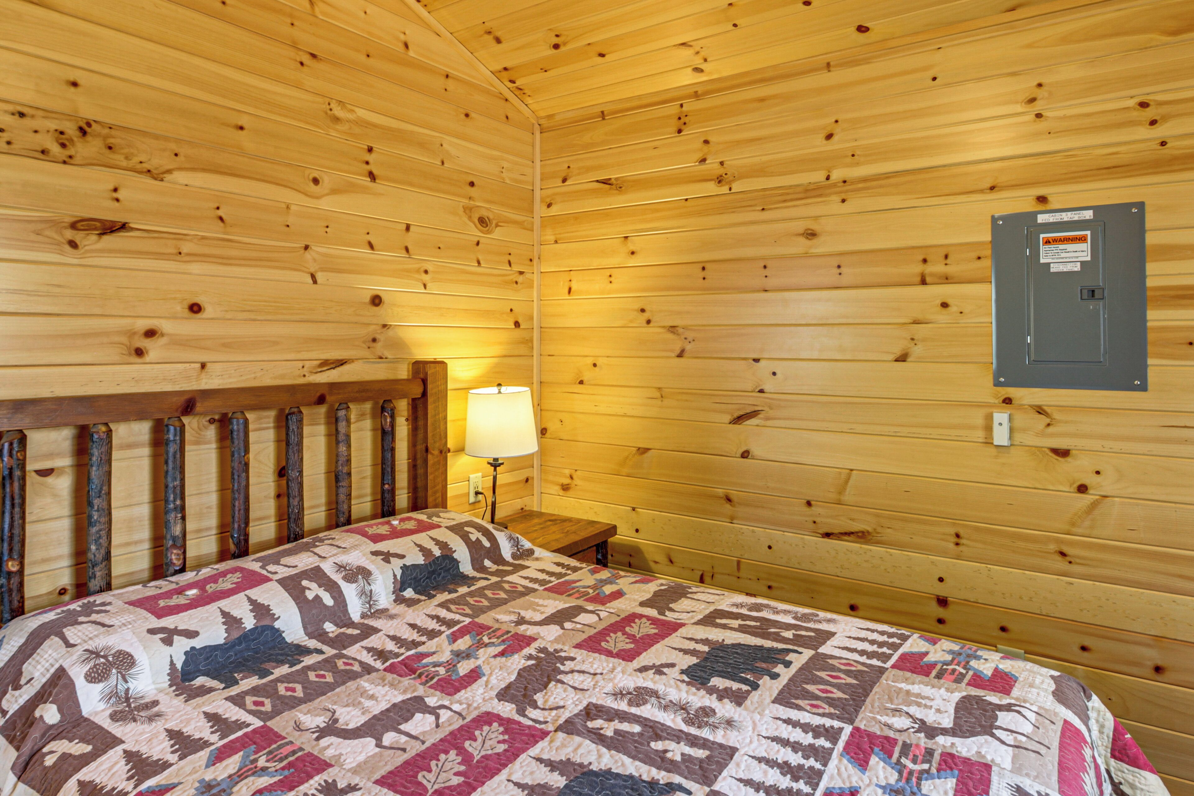 Interior de una cabaña de madera con una cama, una mesita de noche, una lámpara y un cuadro eléctrico de advertencia en la pared.