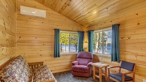 En stuga med träpanel på insidan, inredd med en soffa, två stolar, ett litet bord och ett fönster med gardiner.