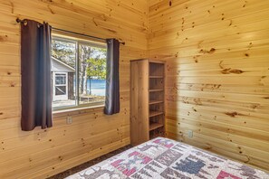 A wooden cabin room with a bed, a window with curtains, and a wooden shelf.