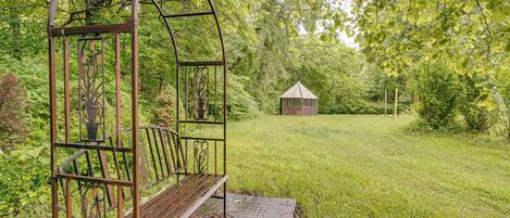 A metal swing structure with a bench, surrounded by lush greenery and a small gazebo in the background.