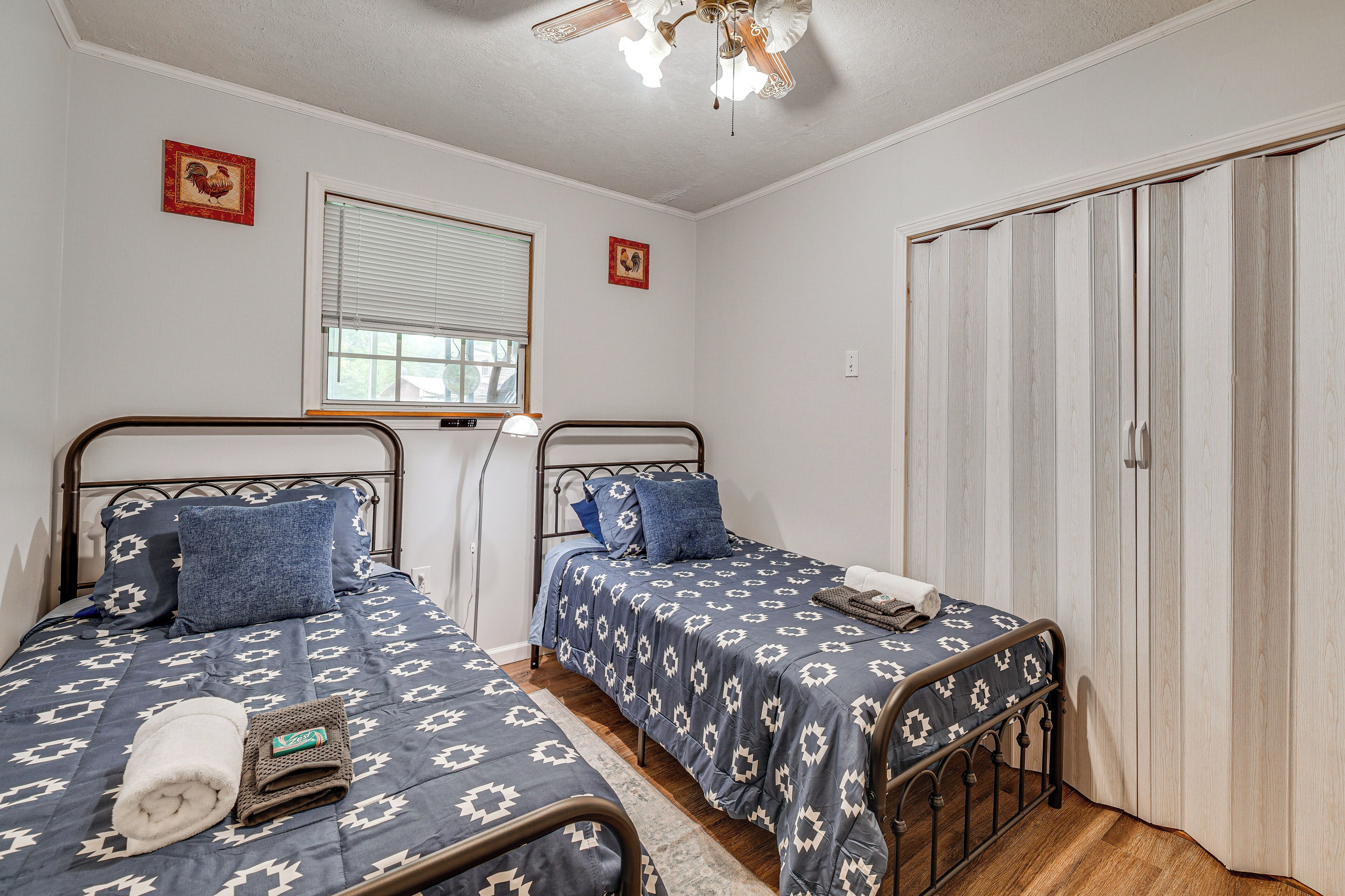 Quarto com duas camas de solteiro, janela com persianas, ventilador de teto e guarda-roupa.
