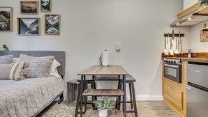 A bedroom with a bed, a small table with a plant, and a kitchen with a stove and refrigerator. - Pet-friendly Quaint Cottage: Park-like Setting (Monroe)