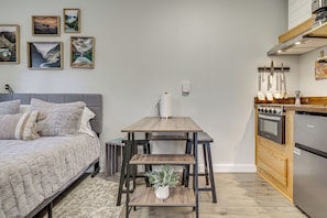 A bedroom with a bed, a small table with a plant, and a kitchen with a stove and refrigerator. - Pet-friendly Quaint Cottage: Park-like Setting (Monroe)