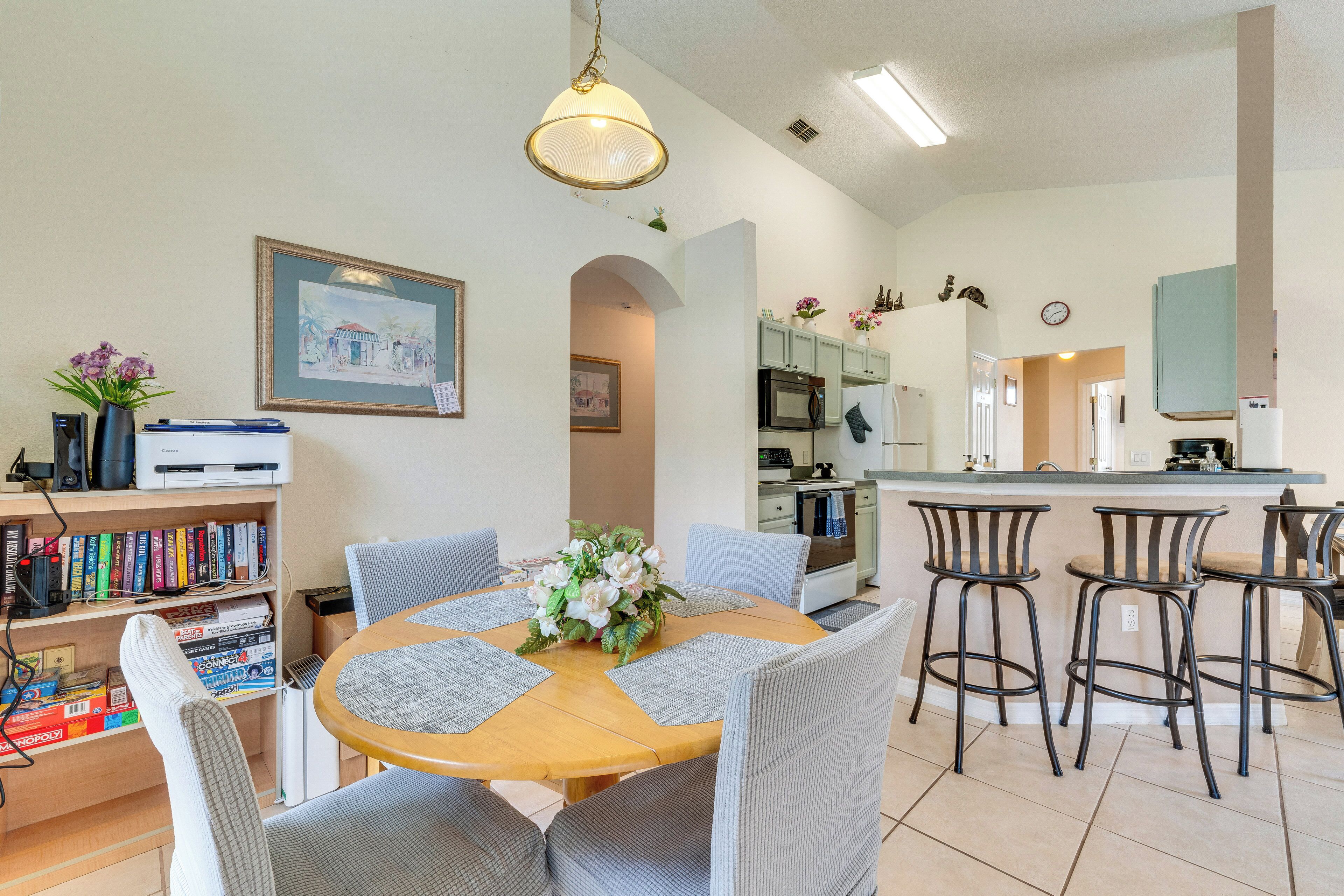 A dining area with a round table, bar stools, a bookshelf, and a kitchen in the background.