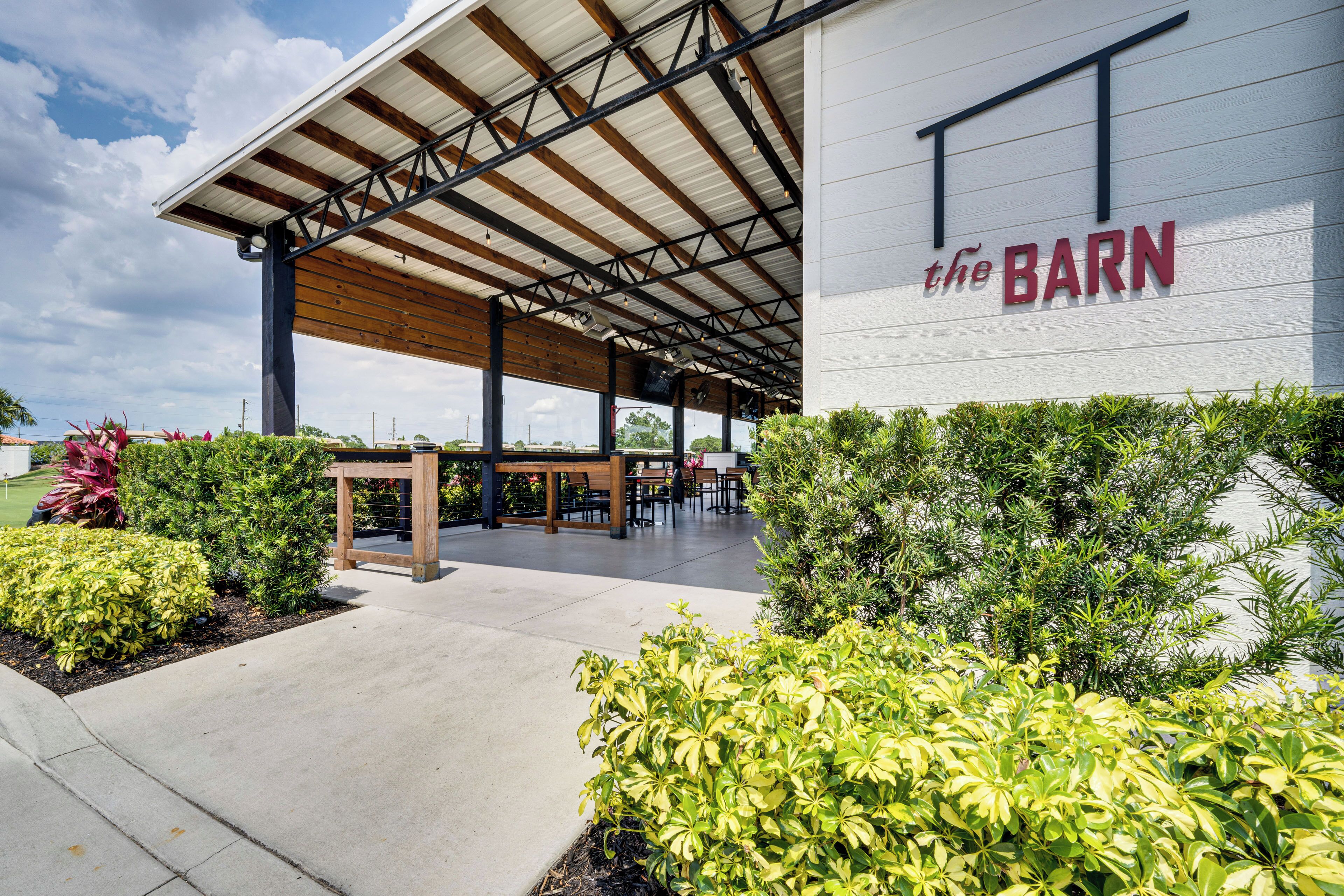 An outdoor seating area with tables and chairs under a covered roof, surrounded by greenery.