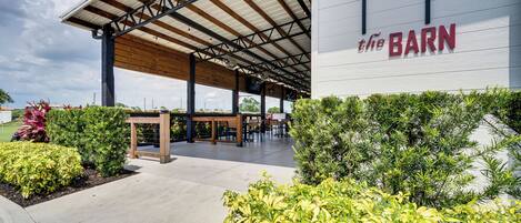 An outdoor seating area with tables and chairs under a covered roof, surrounded by greenery.