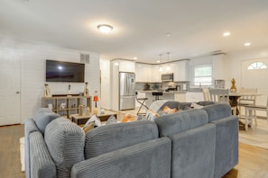 A modern living room with a grey sofa, a flat-screen TV, and a kitchen area with white cabinets and stainless steel appliances.