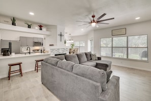 A modern kitchen with grey cabinets, a microwave, and a built-in oven. There is a grey sectional sofa, two wooden stools, and a ceiling fan. - 3 Mi to Seaworld! Cozy Family Home w/ Patio (San Antonio)