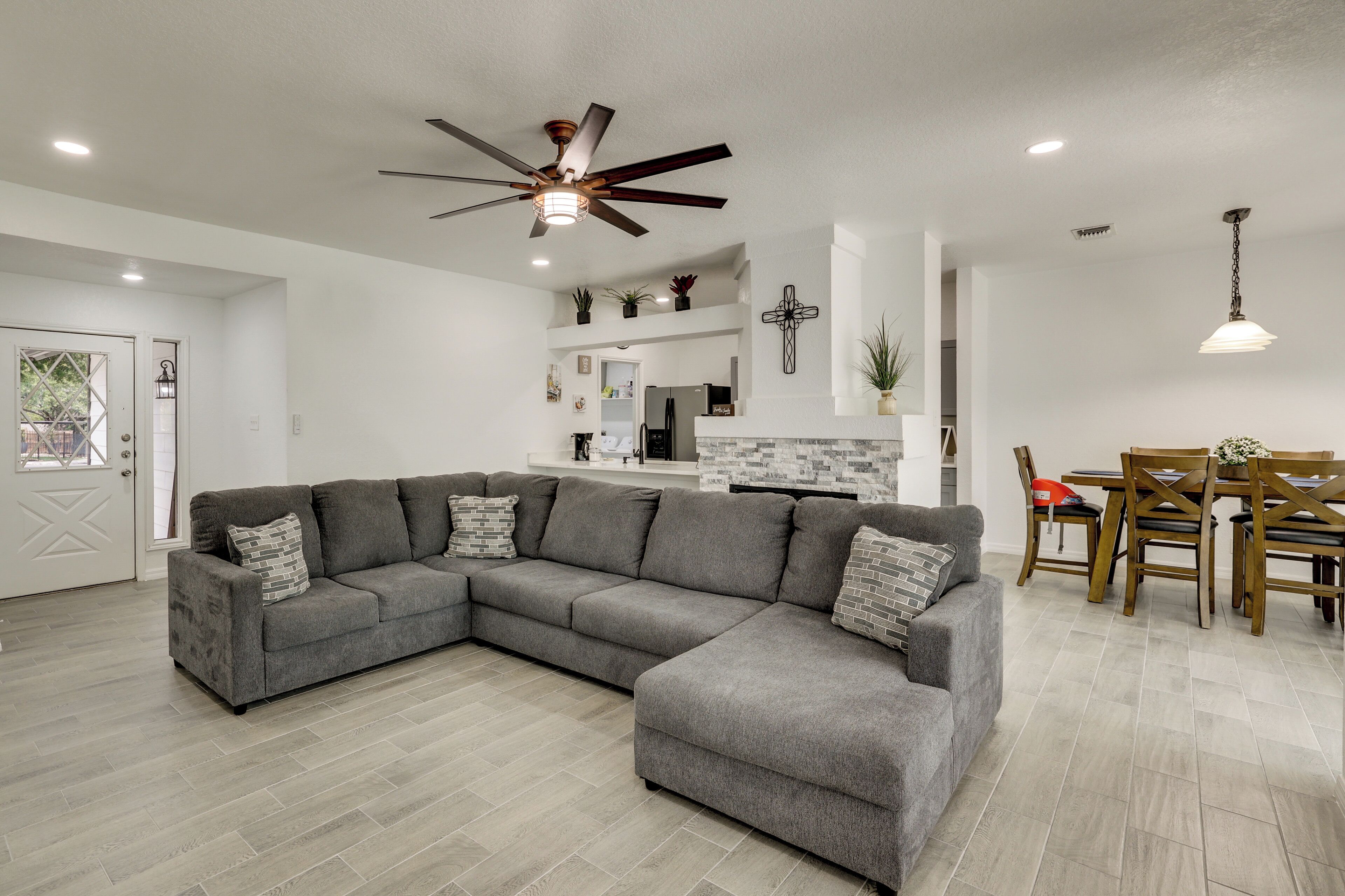A modern living room with a sectional sofa, a dining area with a wooden table and chairs, a ceiling fan, and a cross on the wall.