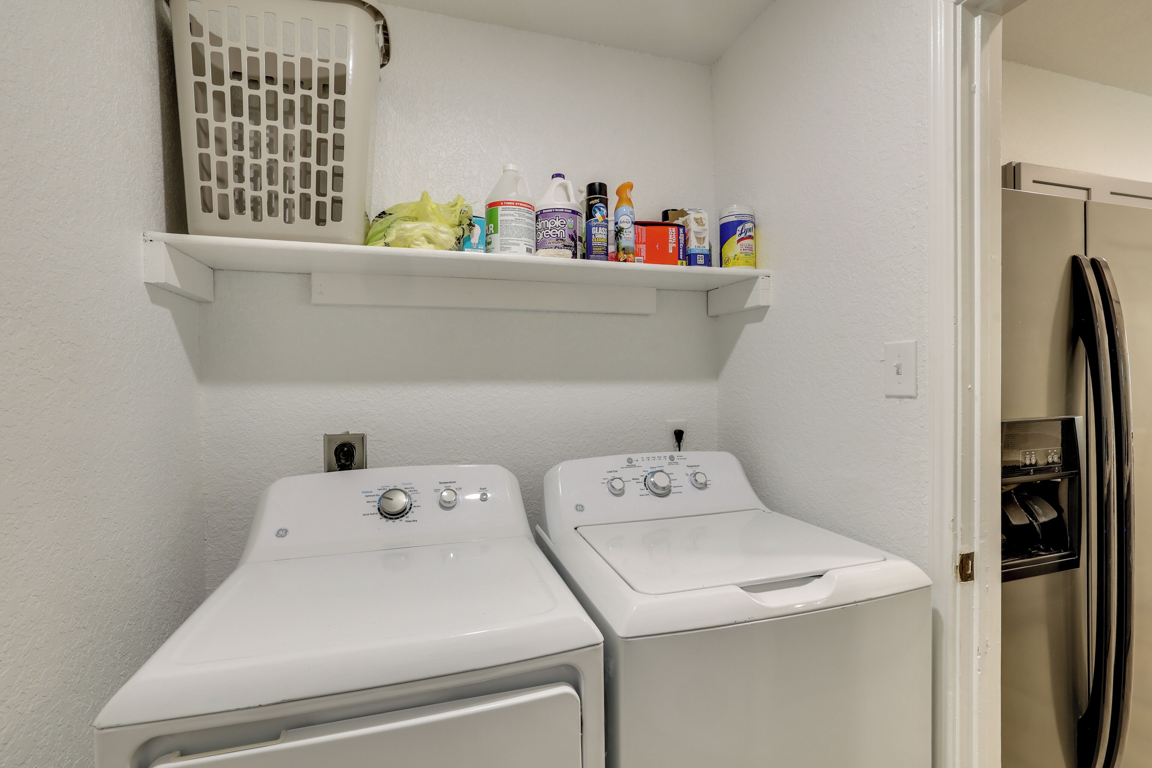 A laundry area with two white washing machines, a shelf with cleaning supplies, and a refrigerator.