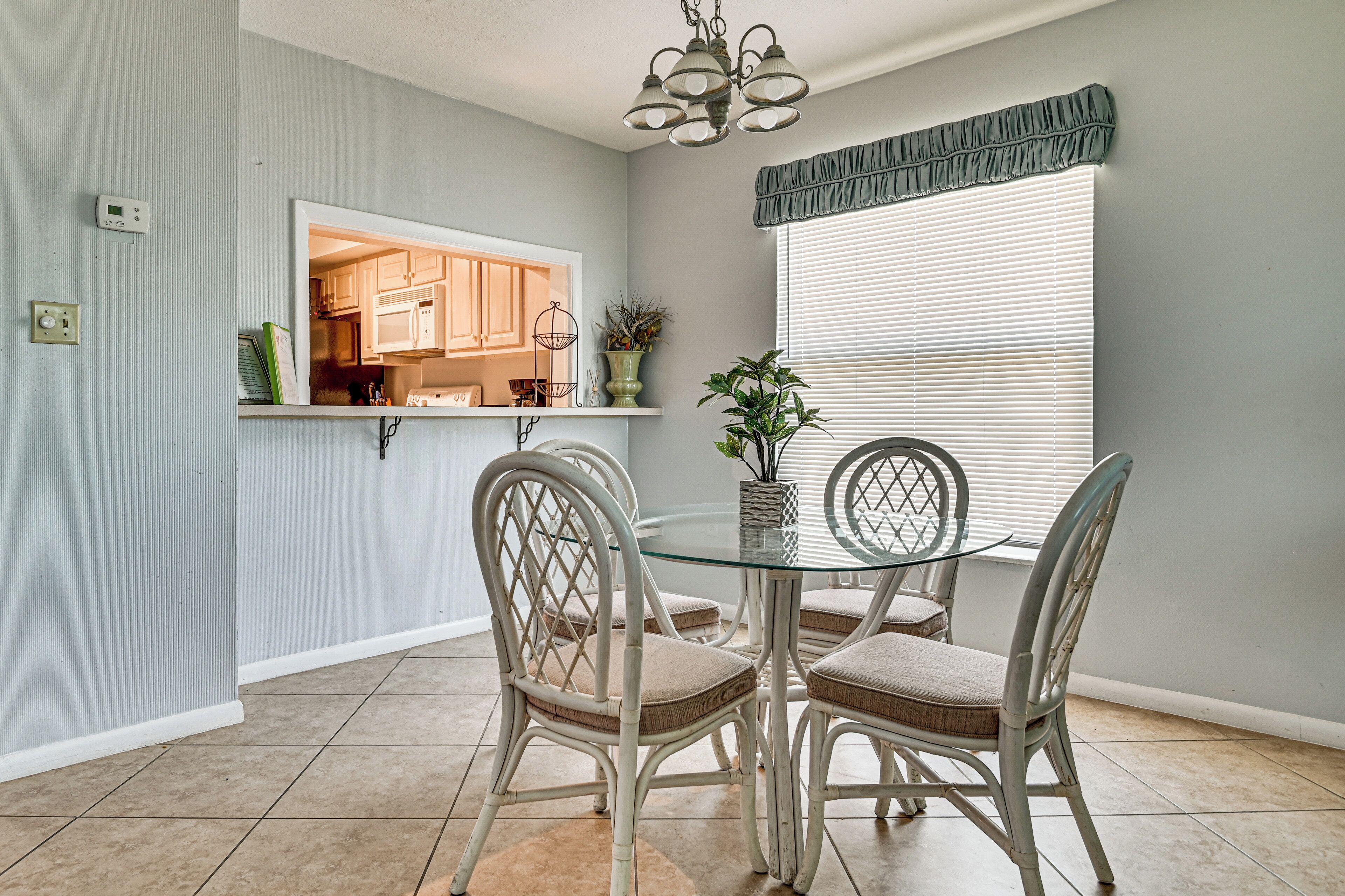 A dining area with a glass table, wicker chairs, a chandelier, and a window with blinds.