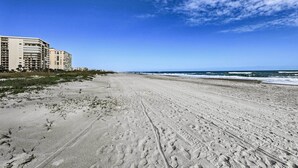 Una amplia playa de arena con huellas, un cielo azul despejado y un edificio al fondo.