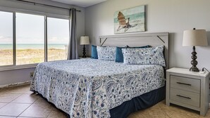 Un dormitorio con un ventanal grande, una cama con ropa de cama estampada, una mesita de noche con una lámpara y un cuadro colgado en la pared.