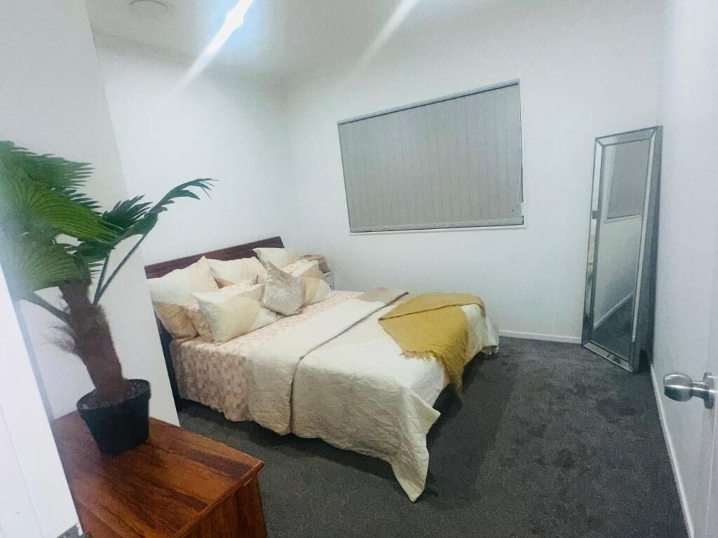 A bedroom with a bed, a potted plant, a bedside table, a window with blinds, and a door.