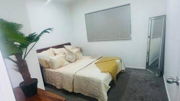 A bedroom with a bed, a potted plant, a bedside table, a window with blinds, and a door.