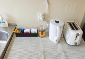 Un coin cuisine comprenant un grille-pain, une bouilloire, un pot de confiture et un flacon de savon liquide, le tout posé sur un tapis à carreaux.