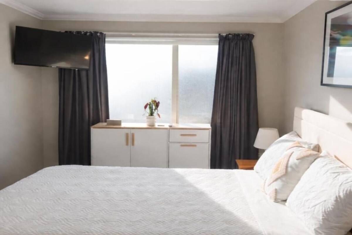 Un dormitorio con cama, televisor montado en la pared, ventana con cortinas, mesita de noche con lámpara y una planta pequeña en el alféizar.