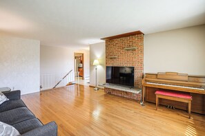 A living room with a wooden floor, a brick fireplace, a television, and a piano. - Extended-stay Home/office, 8 Mi to Dtwn Denver (Lakewood)
