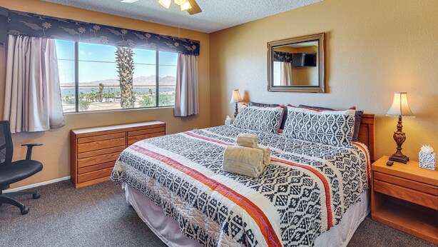 Habitación de hotel con cama, escritorio, silla, mesita de noche, ventilador de techo, ventana con cortinas y vista al exterior.