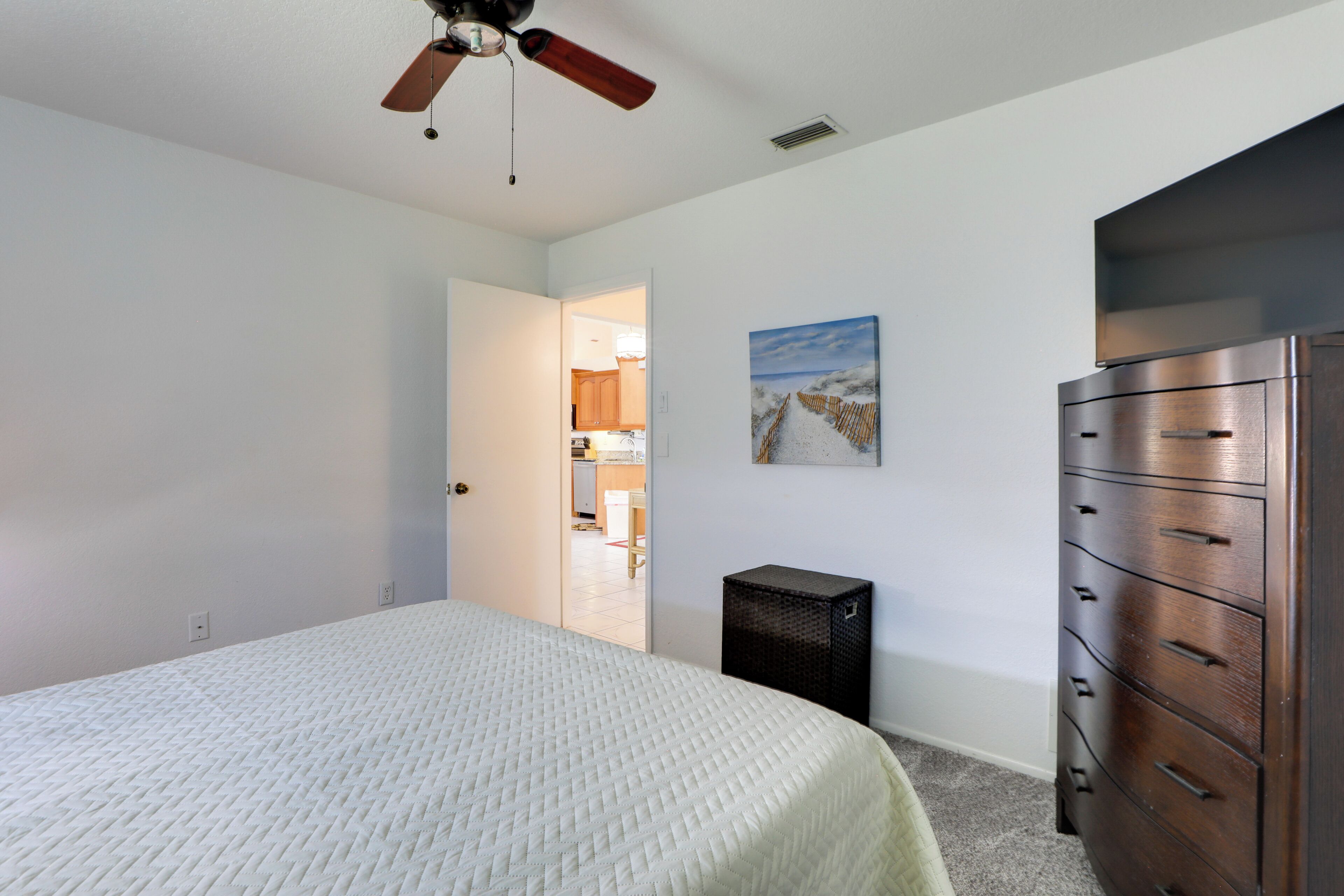 A bedroom with a bed, a dresser, a ceiling fan, a painting, and an open door leading to a kitchen.