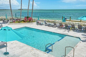 A swimming pool with lounge chairs and umbrellas, surrounded by a stone deck and palm trees, overlooking a body of water. - Beachfront Condo w/ Wide Balcony in Marco Island! (Marco Island)
