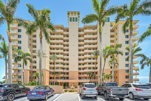 A multi-story hotel with a central courtyard, palm trees, and a parking lot with various vehicles. - Beachfront Condo w/ Wide Balcony in Marco Island! (Marco Island)