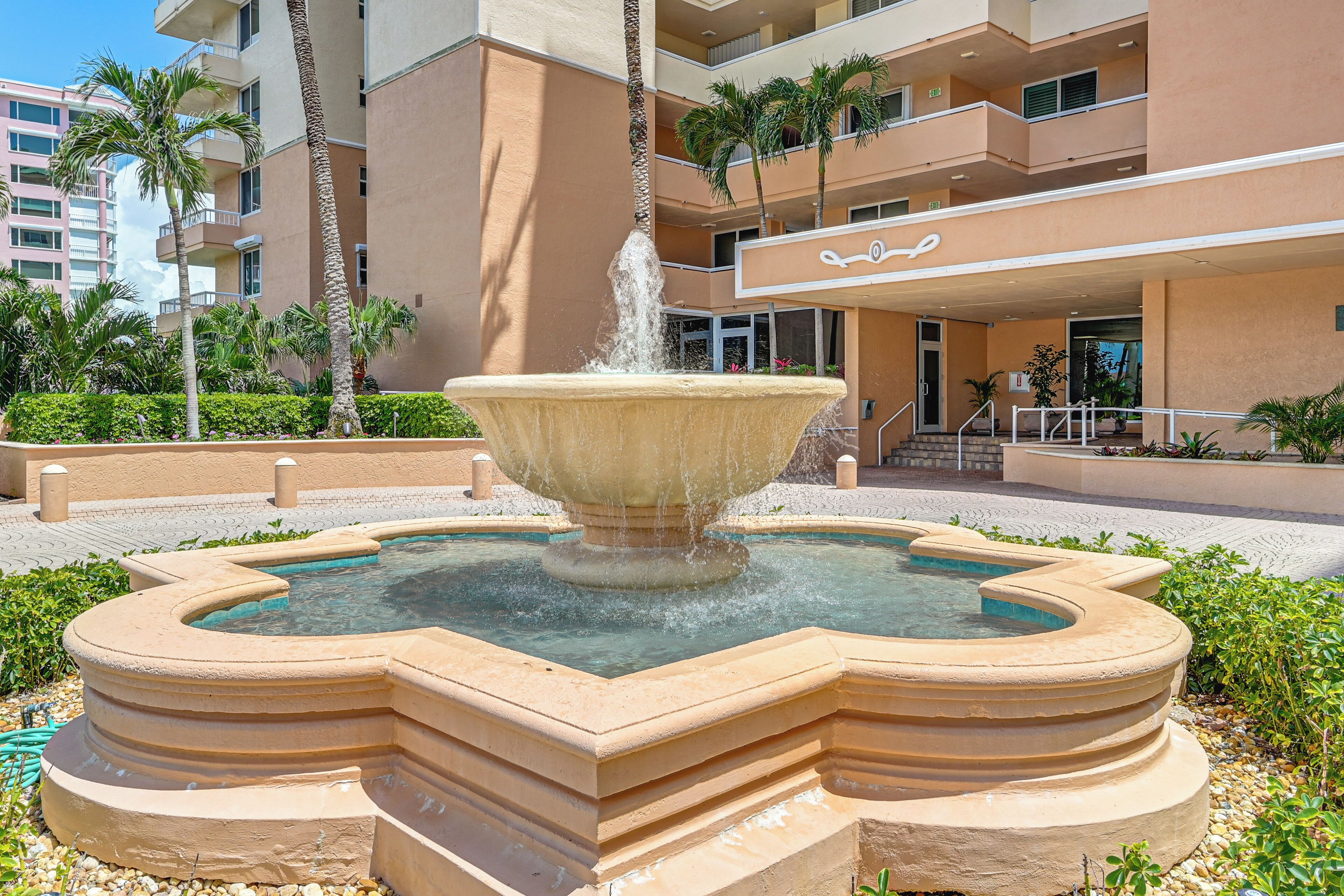 Une fontaine dans une cour intérieure, avec un bassin, entourée de palmiers et d’un bâtiment doté de balcons.