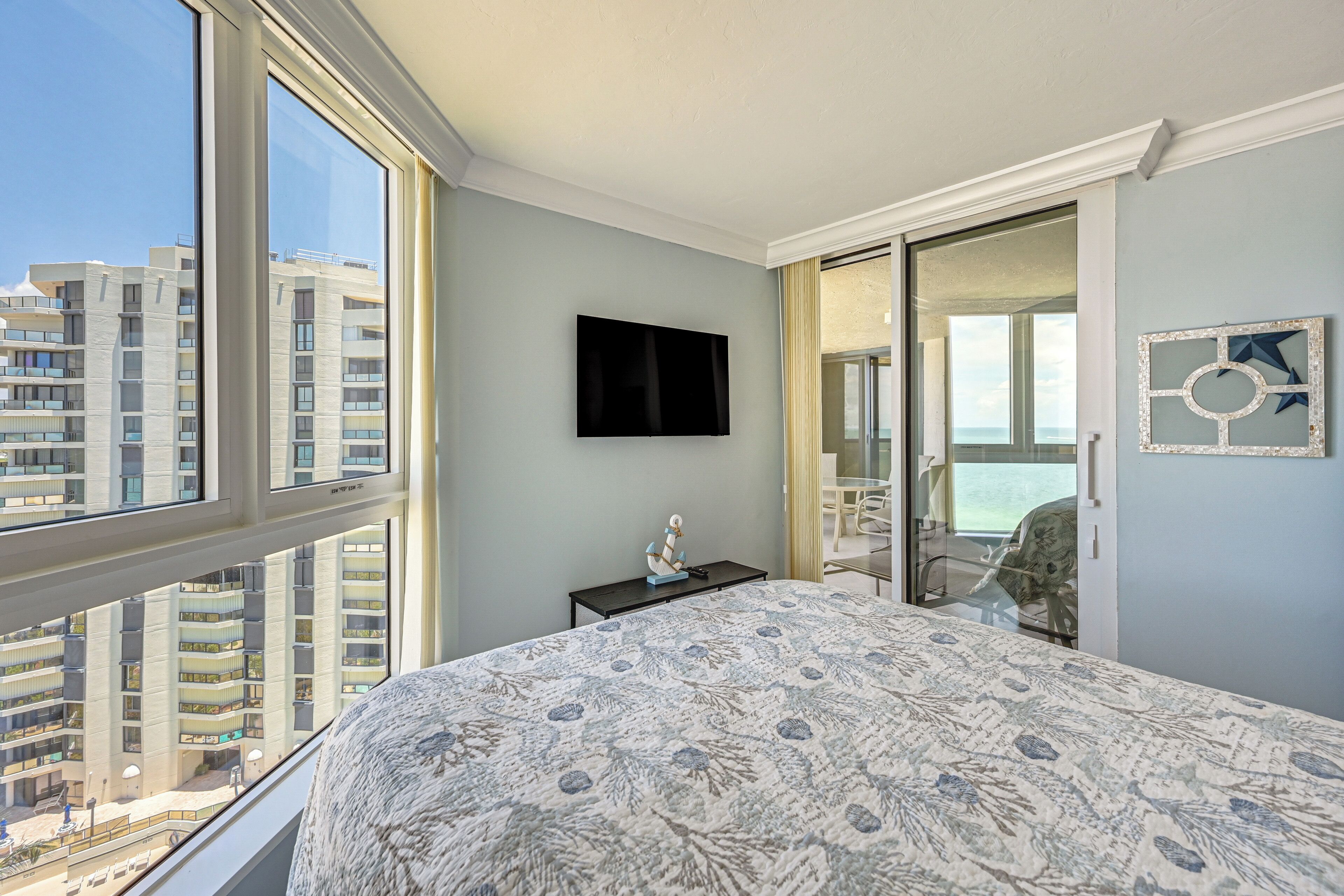 Quarto com uma cama grande, televisão de tela plana, uma mesinha com um objeto decorativo e vista para uma varanda e edifícios.