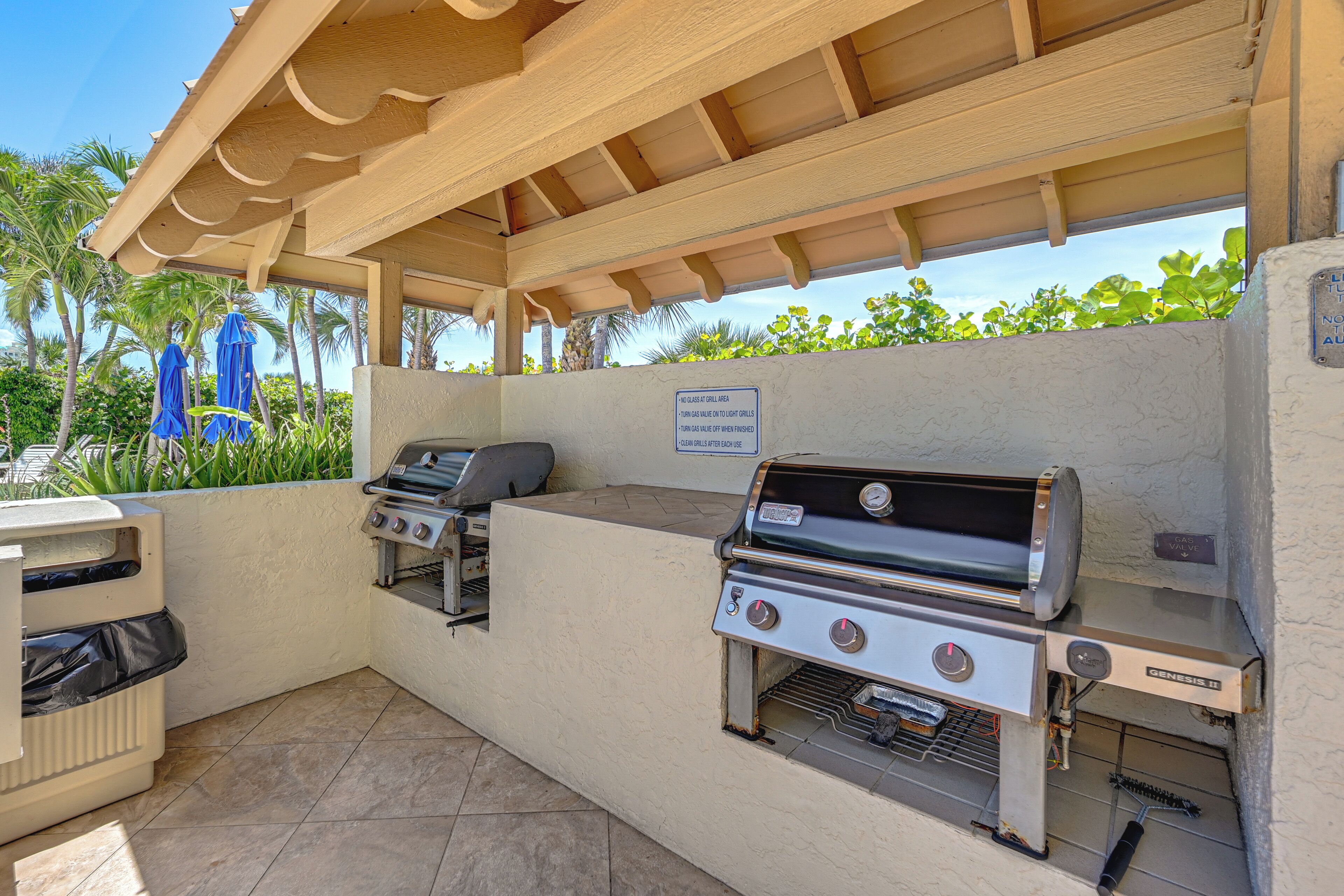 An outdoor barbecue area with a covered roof, two grills, and a trash can.
