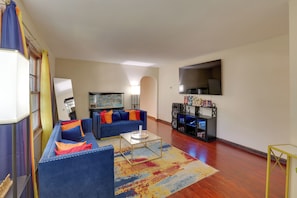 A living room with a sofa set, a coffee table, a flat-screen TV, a bookshelf, and a large window.