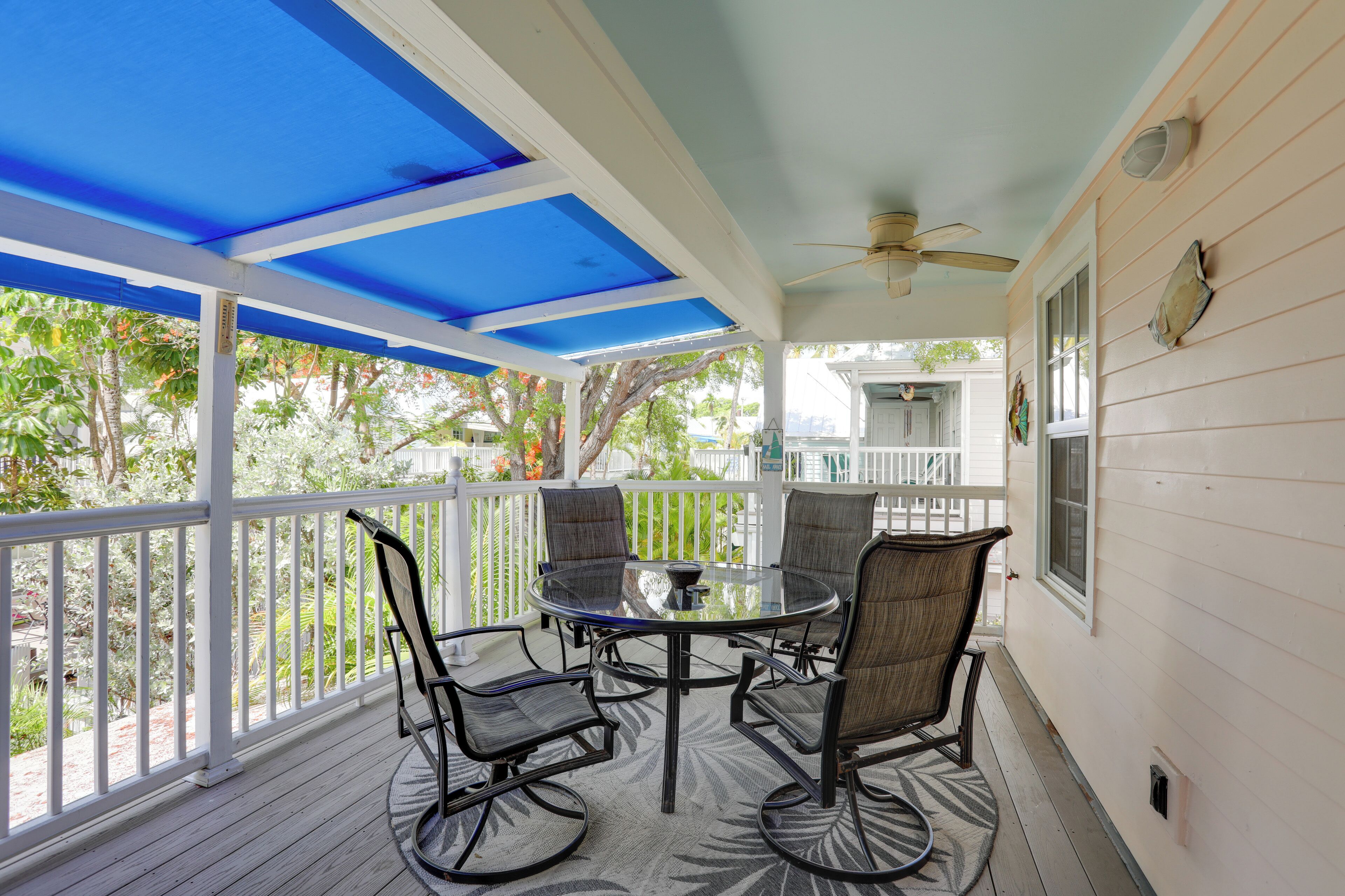A covered outdoor patio with a round table and four chairs, white railings, and a view of greenery.