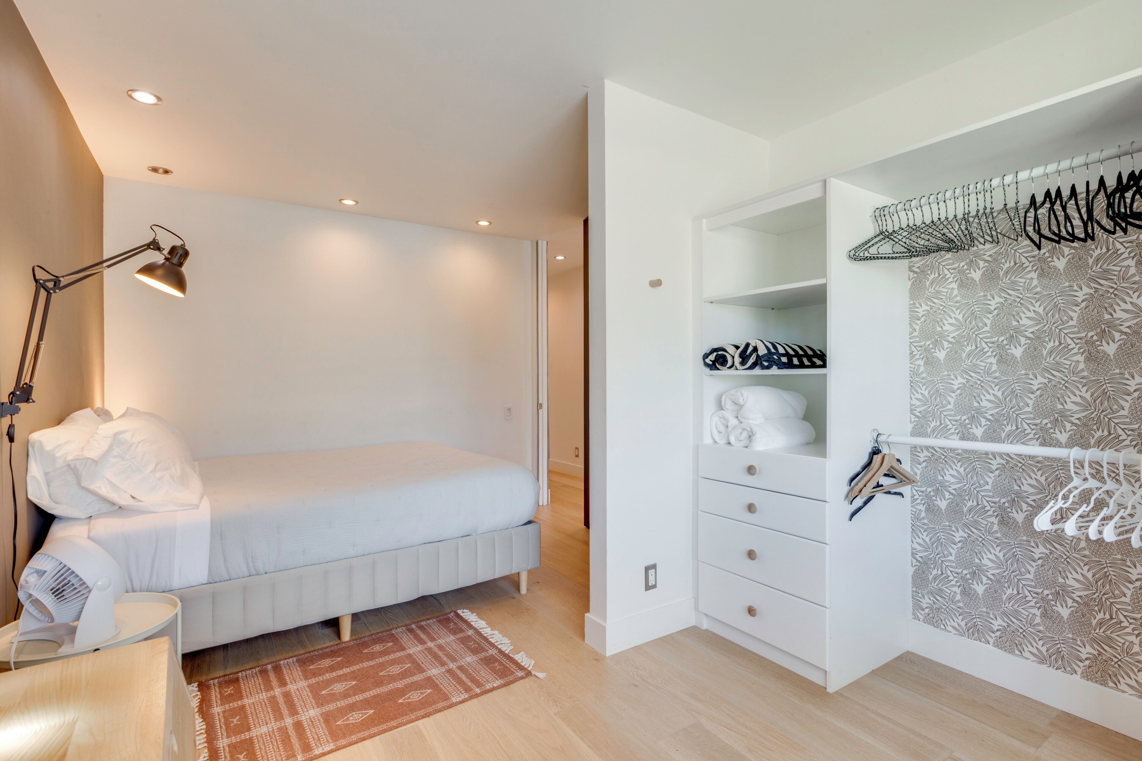 A bedroom with a bed, a bedside table, a lamp, a wardrobe, and a hanging rack.