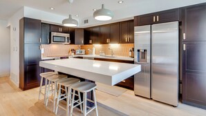 A modern kitchen with dark cabinets, stainless steel appliances, a large island with stools, and pendant lighting. - Unwind Poolside, Walk to El Paseo: Desert Oasis! (Palm Desert)