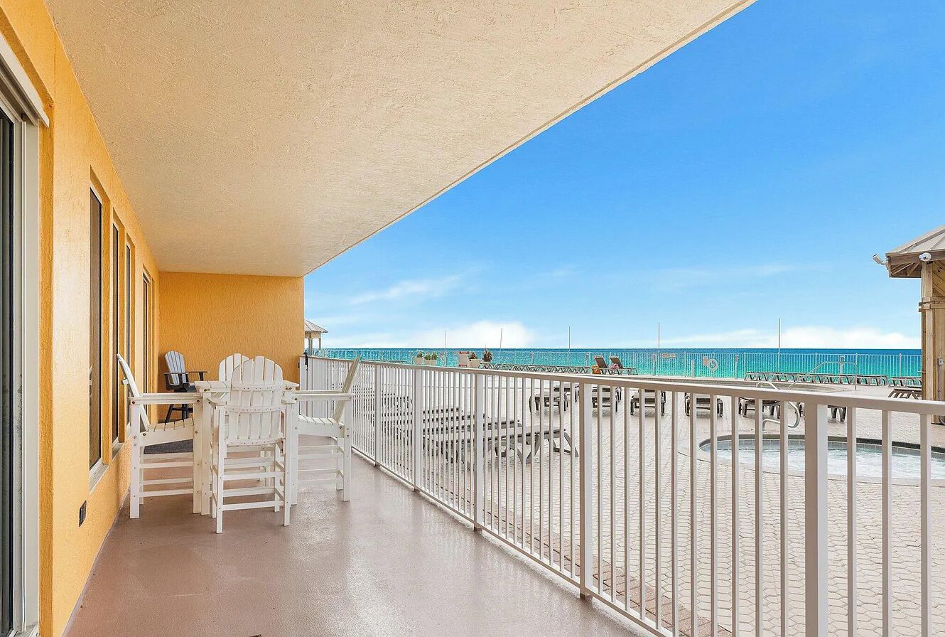 A balcony with a view of the beach, white chairs, and a railing.
