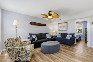 Ein Wohnzimmer mit dunkelblauer Sofagarnitur, einem gemusterten Sessel, einem runden Hocker, einem Deckenventilator und einem an der Wand befestigten Schild.