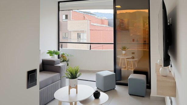 Um espaço de estar moderno com sofá, uma pequena mesa redonda, uma planta em vaso e vista para edifícios através de uma grande janela.