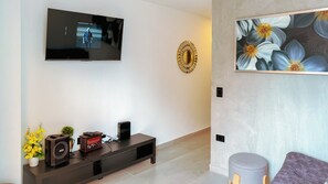 Een moderne woonkamer met een flatscreen televisie aan de muur, een donker houten entertainmentkast met speakers en een decoratief object, een ronde spiegel met gouden lijst, en een groot schilderij met bloemen aan de muur.