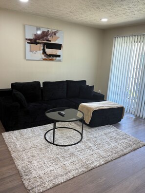 A living room with a black sofa, a glass coffee table, a beige rug, and a painting on the wall.