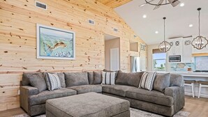 A modern living room with a sectional sofa, a matching ottoman, a wooden wall with a picture, and a kitchen area with white cabinets and a TV.