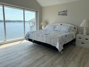 Een slaapkamer met een groot raam, een bed met een beddeken met een patroon, een nachtkastje en een wanddecoratie.