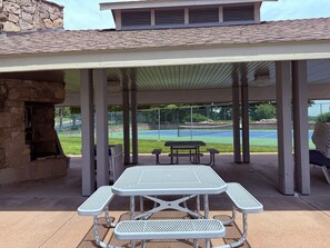 Un patio couvert, équipé d’une table et de bancs, donnant sur un court de tennis.