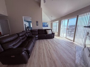 Een ruime woonkamer met een grote hoekbank, een eethoek met stoelen en uitzicht door grote ramen.